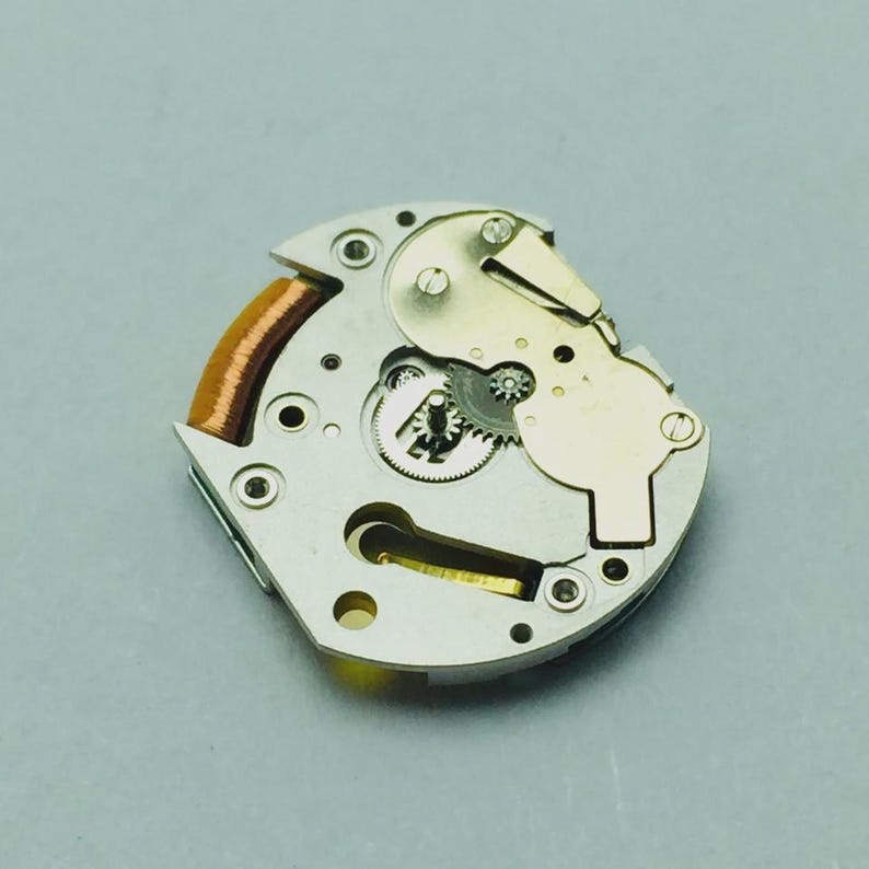 May include: A detailed view of a watch movement, showcasing delicate gears, a copper coil, and metallic parts. The movement is silver and gold, with tiny screws and a complex design. The background is a plain grey.