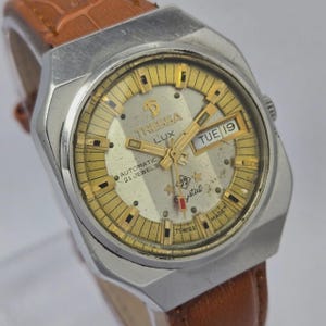 Pode incluir: Relógio automático Tressa Lux vintage com caixa prateada e mostrador dourado. O relógio apresenta exibição de dia e data, 21 rubis e uma bracelete de couro castanho. O mostrador tem o texto "Tressa Lux" e "Crystal".