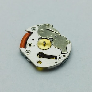May include: Close-up of a watch movement with a gold-colored gear at the center. The movement has silver-colored plates and a red and orange component on the left side. The image is taken from a top-down perspective.