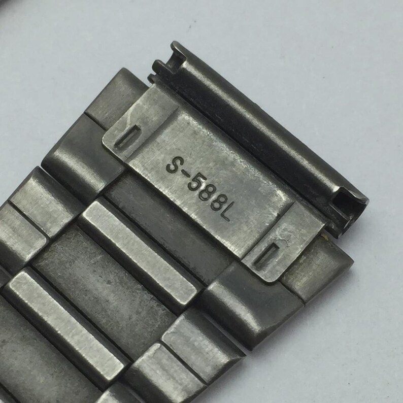Puede incluir: Primer plano de una correa de reloj plateada con el texto "S-588L" grabado en una placa rectangular. La correa est&aacute; compuesta por m&uacute;ltiples eslabones rectangulares.