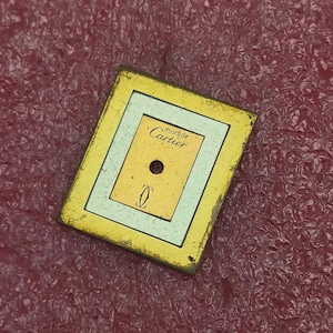 May include: A rectangular watch dial featuring a yellow border, a white inner frame, and a central yellow panel. The text 'must de Cartier' is printed in black script above a central circular hole, with the Cartier double C logo positioned below.