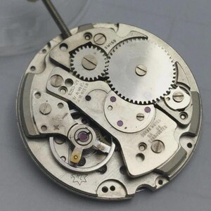 May include: Close-up of a watch movement with gears, screws, and other mechanical components. The watch mechanism is silver-toned with visible text that reads "SWISS" and "SORNA WATCH SA DE HAND-ADJUSTED".