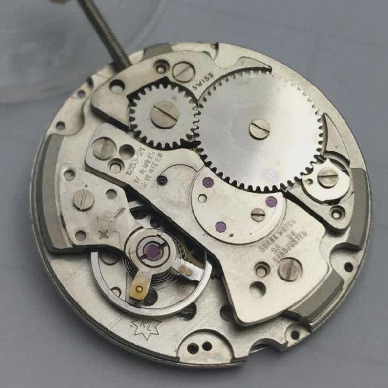 May include: Close-up of a watch movement with gears, screws, and other mechanical components. The watch mechanism is silver-toned with visible text that reads "SWISS" and "SORNA WATCH SA DE HAND-ADJUSTED".