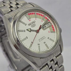 Pu&ograve; includere: Un orologio da polso automatico Seiko in acciaio inossidabile con quadrante bianco. L'orologio presenta un display giorno e data, dettagli rossi e un cinturino argentato. Il quadrante mostra il testo "SEIKO" e "AUTOMATIC 21 JEWELS".