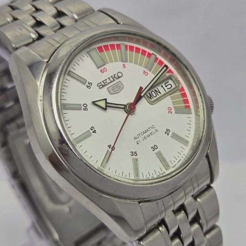 Pu&ograve; includere: Un orologio da polso automatico Seiko in acciaio inossidabile con quadrante bianco. L'orologio presenta un display giorno e data, dettagli rossi e un cinturino argentato. Il quadrante mostra il testo "SEIKO" e "AUTOMATIC 21 JEWELS".