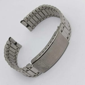 Puede incluir: Una correa de reloj de acero inoxidable plateado con un cierre rectangular. La correa está compuesta por pequeños eslabones rectangulares que se expanden y contraen. El cierre tiene un acabado cepillado y un pequeño emblema grabado.