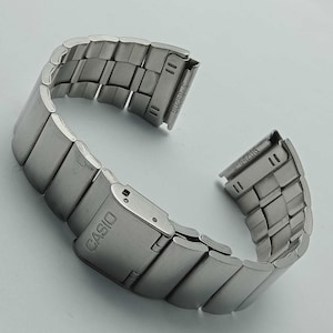 May include: A silver-coloured metal watch strap with a rectangular link design. The strap features the brand name "CASIO" on the clasp. The strap is curved and appears to be new.