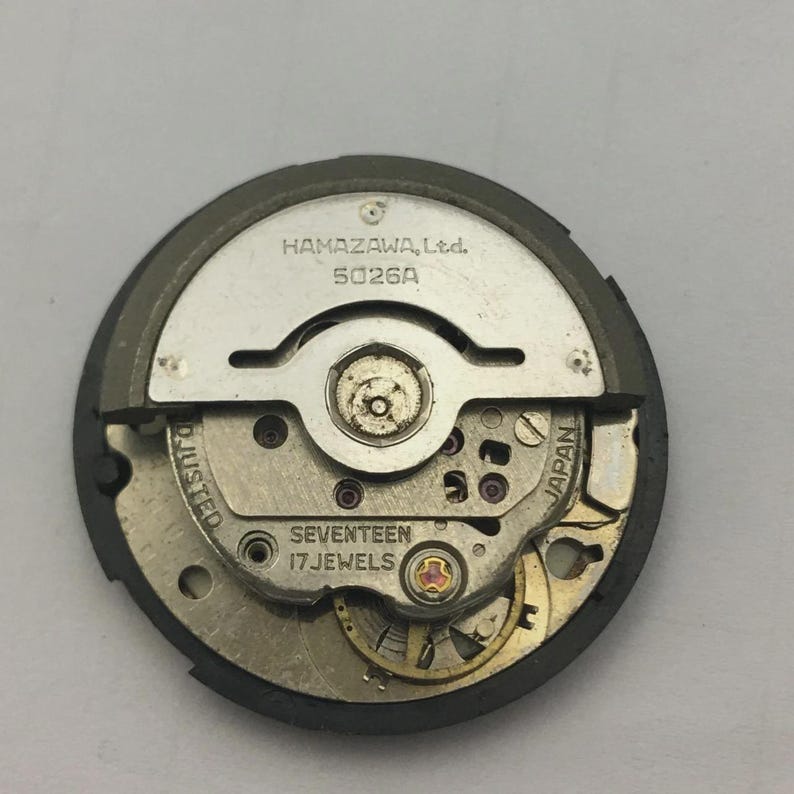 Może przedstawiać: Zbliżenie mechanizmu zegarka z napisem "HAMAZAWA, Ltd. 5026A" i "SEVENTEEN 17 JEWELS". Mechanizm zegarka ma srebrno-złotą kolorystykę, z widocznym słowem "JAPAN".