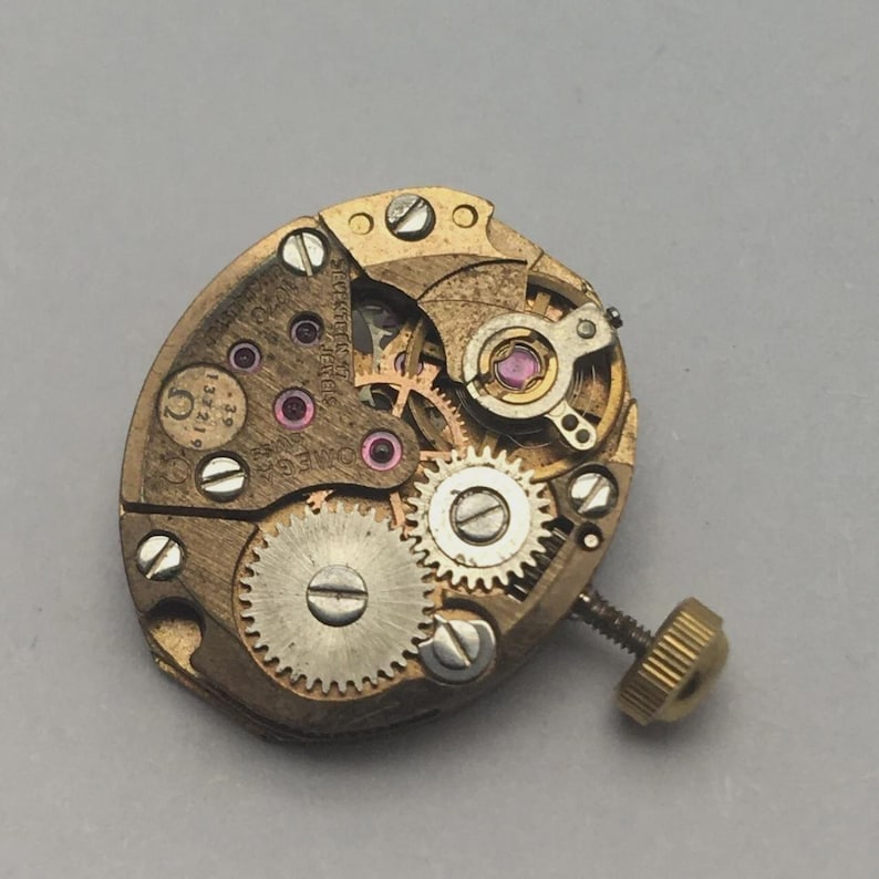 Pode incluir: Close-up de um movimento de rel&oacute;gio Omega vintage. O mecanismo apresenta in&uacute;meras engrenagens pequenas, parafusos e joias em tons de ouro e rosa. O movimento do rel&oacute;gio tem formato oval, com haste de corda e coroa.