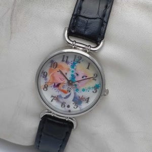以下が含まれることがあります： 黒い革バンドが付いたシルバーカラーの腕時計。文字盤には、エルサ、アナ、オラフなど、映画「アナと雪の女王」のキャラクターがカラフルに描かれています。丸い文字盤にはアラビア数字が使われています。