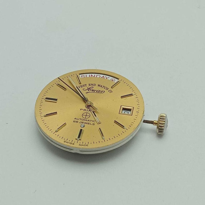 May include: A gold-toned watch face with the text "West End Watch Co Sowar" and "Automatic 25 Jewels". The watch face has gold hands and markers, a date window, and a crown. The word "Sunday" is visible.