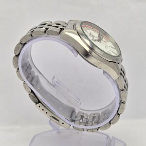 Pu&ograve; includere: Un orologio argentato con quadrante bianco e dettagli rossi. L'orologio ha un cinturino in metallo e un quadrante rotondo. L'orologio &egrave; esposto su un supporto di plastica trasparente.