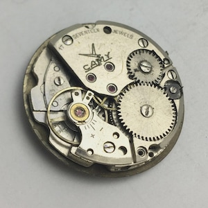 K&ouml;nnte beinhalten: Nahaufnahme eines Vintage-Uhrwerks mit sichtbaren Zahnr&auml;dern, Federn und Juwelen. Der silberfarbene Mechanismus tr&auml;gt den Markennamen "CAMY" und den Text "SEVENTEEN JEWELS". Die Aufnahme zeigt die filigrane Konstruktion des Uhrwerks.