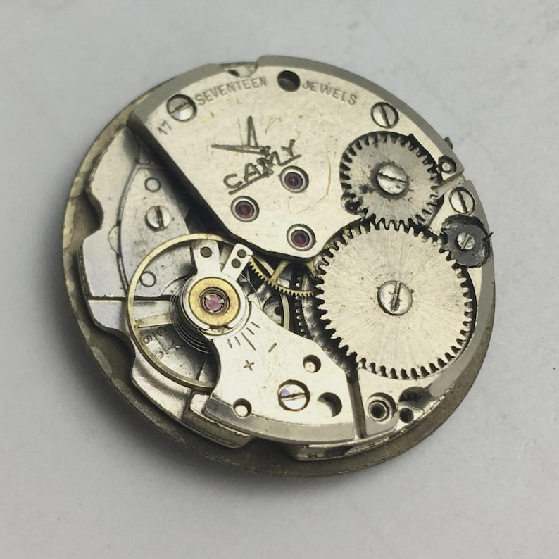 K&ouml;nnte beinhalten: Nahaufnahme eines Vintage-Uhrwerks mit sichtbaren Zahnr&auml;dern, Federn und Juwelen. Der silberfarbene Mechanismus tr&auml;gt den Markennamen "CAMY" und den Text "SEVENTEEN JEWELS". Die Aufnahme zeigt die filigrane Konstruktion des Uhrwerks.