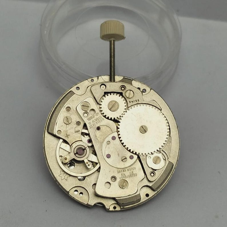 May include: A disassembled watch movement with visible gears, jewels, and a winding stem. The metal components are primarily gold-toned, with the word "SWISS" visible. The watch movement is a complex mechanism, showcasing the intricate inner workings of a timepiece.