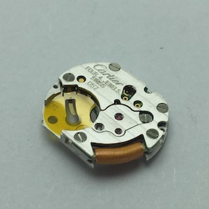Pu&ograve; includere: Primo piano di un movimento di orologio Cartier. Il meccanismo argentato presenta il marchio "Cartier" e il testo "FOUR 4 JEWELS SWISS 057". Il movimento ha un componente dorato e una bobina marrone.
