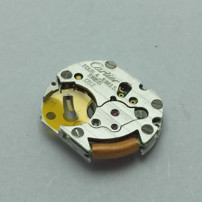 Pu&ograve; includere: Primo piano di un movimento di orologio Cartier. Il meccanismo argentato presenta il marchio "Cartier" e il testo "FOUR 4 JEWELS SWISS 057". Il movimento ha un componente dorato e una bobina marrone.