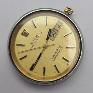 Op de afbeelding: Een vintage Omega Seamaster Chronometer wijzerplaat met een goudkleurige wijzerplaat. De horlogewijzerplaat toont de dag en datum, met "ZA 6" zichtbaar. Het heeft een zilverkleurige lunette en een goudkleurige kroon. De wijzerplaat bevat "OMEGA MEGASSONIC 720 Hz" en "Seamaster CHRONOMETER".