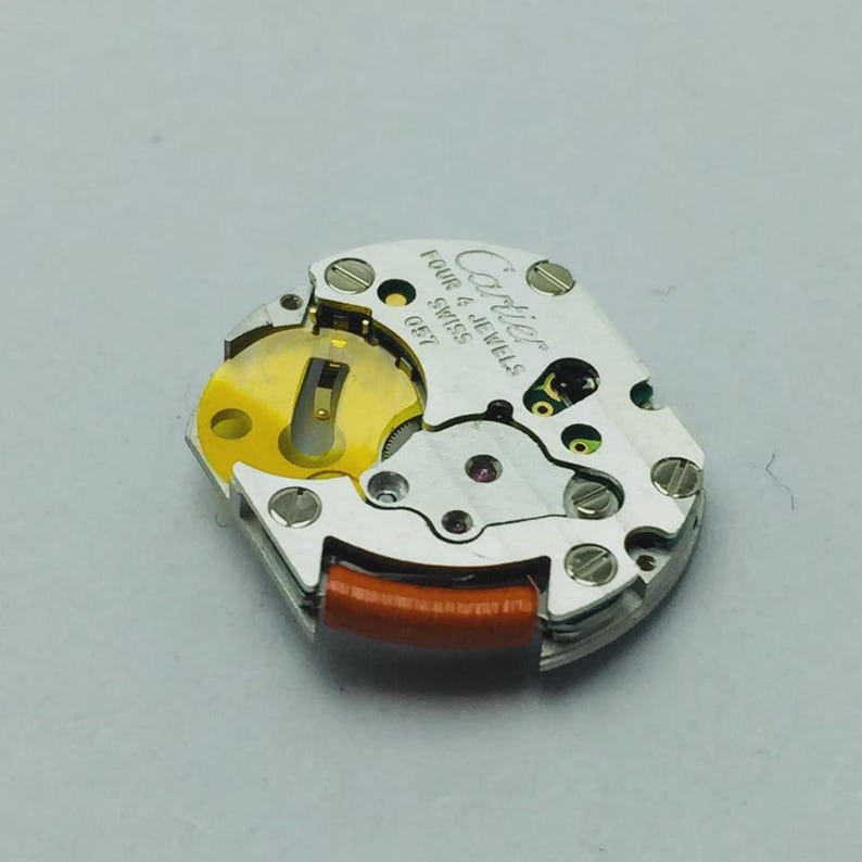 May include: A close-up of a Cartier watch movement. The silver-toned mechanism features the text "Cartier Four 4 Jewels Swiss GE7". The movement has a gold-colored rotor, red and black components, and small screws.