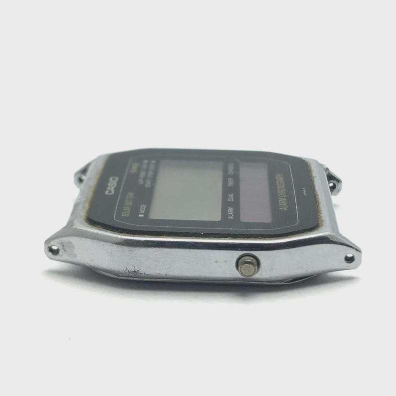 May include: A silver-toned Casio digital watch with a black display. The watch features a rectangular face with buttons and text indicating functions like "Alarm Chronograph" and "Stop Battery". The watch has a classic, retro design.