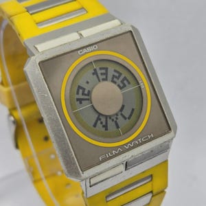 May include: A vintage Casio Film Watch with a yellow and silver band. The watch face is rectangular with a digital display showing the time. The watch face has a gray background with a yellow circular design and the text "FILM WATCH".