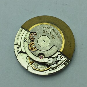 Puede incluir: Primer plano de un movimiento de reloj con engranajes, joyas y un rotor dorado visibles. El mecanismo del reloj est&aacute; marcado como "Revue Swiss 21 Jewels". Los intrincados detalles de las piezas del reloj son claros.