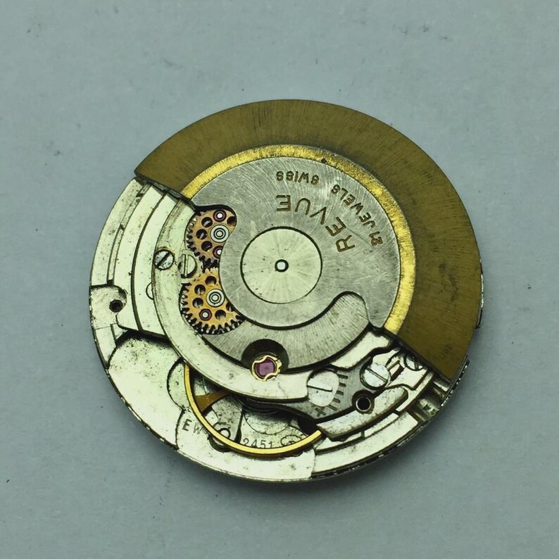 Puede incluir: Primer plano de un movimiento de reloj con engranajes, joyas y un rotor dorado visibles. El mecanismo del reloj est&aacute; marcado como "Revue Swiss 21 Jewels". Los intrincados detalles de las piezas del reloj son claros.