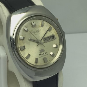 Op de afbeelding: Een vintage zilverkleurig horloge met een zwarte leren band. De wijzerplaat is lichtgoudkleurig met rechthoekige uurmarkeringen en de dag van de week wordt weergegeven. De wijzerplaat leest "Spacer Waterproof".