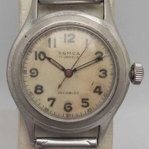 Puede incluir: Reloj de pulsera SEMCA vintage con caja plateada y esfera color crema. El reloj presenta números negros, manecillas y el texto "SEMCA 17 JEWELS" e "INCABLOC". Cuenta con una correa extensible de metal.