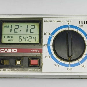 Op de afbeelding: Een vintage Casio HT-100 timer met een digitale display die 12:12 AM en een timer van 64:24 weergeeft. Het apparaat heeft een witte en zilveren behuizing, een blauwe wijzerplaat en rode accenten. Het heeft knoppen voor start/stop en minuten selectie.