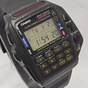Puede incluir: Un reloj Casio negro con mando a distancia de mu&ntilde;eca con calculadora y funciones de alarma. El reloj muestra la hora y el d&iacute;a. Tiene un teclado con botones para varias funciones, incluido el control de TV. La correa del reloj es negra.