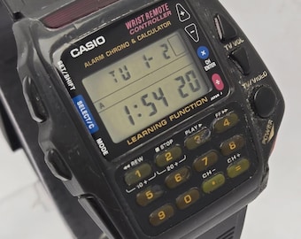 May include: A black Casio wrist remote controller watch with a calculator and alarm functions. The watch displays the time and day. It has a keypad with buttons for various functions, including TV control. The watch band is black.