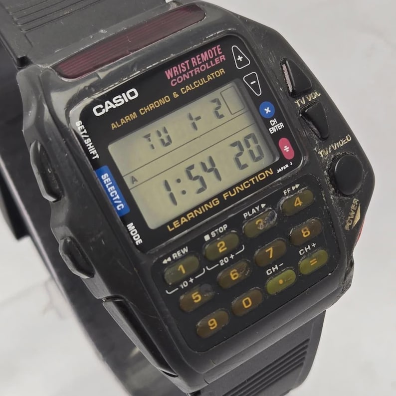 Puede incluir: Un reloj Casio negro con mando a distancia de mu&ntilde;eca con calculadora y funciones de alarma. El reloj muestra la hora y el d&iacute;a. Tiene un teclado con botones para varias funciones, incluido el control de TV. La correa del reloj es negra.