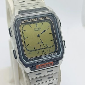 Puede incluir: Un reloj Citizen Quartz plateado con una esfera rectangular. La esfera es dorada con detalles en negro y muestra la fecha 4.15 TU. El reloj tiene una correa plateada y presenta el texto "ALARM CHRONO DUAL TIME".