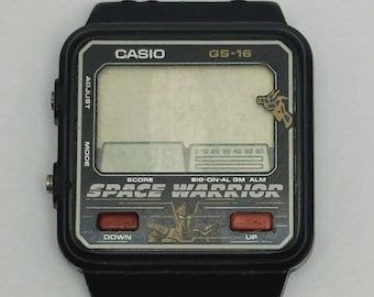 May include: Black Casio GS-16 Space Warrior digital watch. The watch face displays the text "SPACE WARRIOR" and has red "DOWN" and "UP" buttons. The watch has a black strap and a small graphic of a robot.