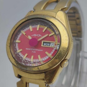 May include: A gold-coloured AKA watch with a red face and a date display showing "MON 22". The watch has gold hands and markers, and the face includes the text "WATER 10 BAR RESIST ALBA".