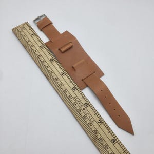 Könnte beinhalten: Ein hellbraunes Lederarmband mit silberner Schnalle. Das Armband hat eine breite Basis mit zwei kleinen Schlaufen und einen schmaleren Riemen mit mehreren Löchern. Ein Holzlineal mit cm-Markierungen liegt daneben.