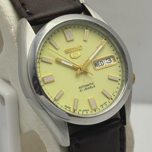 May include: A silver-toned Seiko 5 automatic wristwatch with a light yellow face, gold hands, and a brown leather strap. The watch face displays the day and date, with the text "AUTOMATIC 21 JEWELS" and "MADE IN JAPAN".