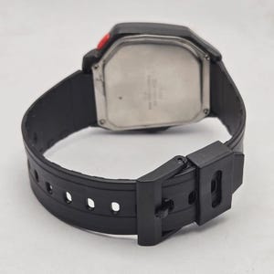 Puede incluir: Reloj digital negro con esfera rectangular y correa de goma negra. El reloj tiene una parte trasera plateada y un bot&oacute;n rojo. La correa se asegura con una hebilla negra.