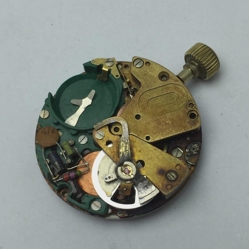 May include: Close-up of a disassembled watch movement. The mechanism features brass-colored plates, a green component, and various small parts. A gold-colored crown is visible on the right side. The watch movement is set against a gray background.