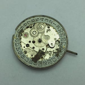 May include: A close-up of a watch movement with visible gears, a date dial, and a small stem. The metal components are silver and show signs of wear. The date dial is marked with numbers from 1 to 31.