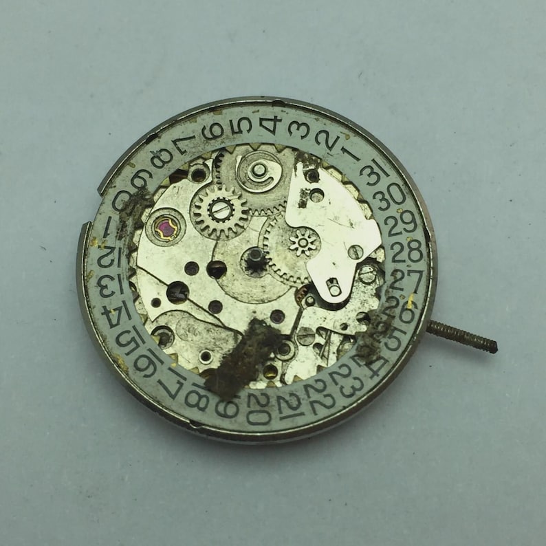 May include: A close-up of a watch movement with visible gears, a date dial, and a small stem. The metal components are silver and show signs of wear. The date dial is marked with numbers from 1 to 31.