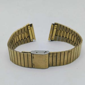 Puede incluir: Una correa de reloj Casio dorada, con un diseño de eslabones rectangulares. La correa tiene un cierre con el logotipo de Casio. La correa está diseñada para expandirse y contraerse. La correa se muestra sobre un fondo blanco.