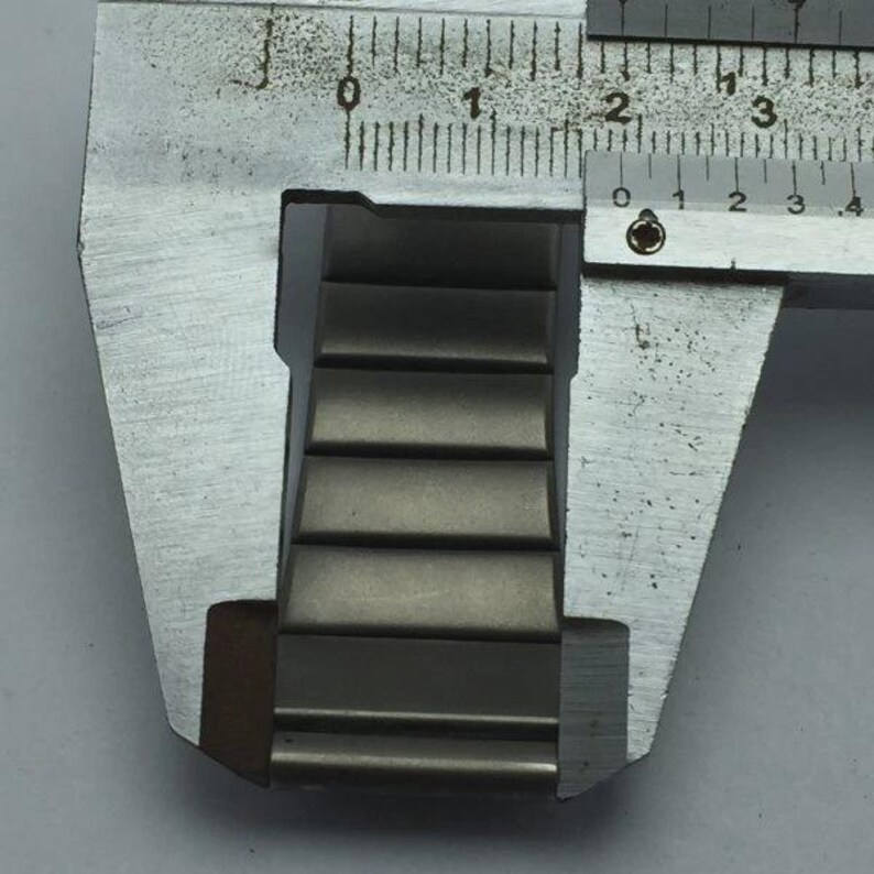 Puede incluir: Primer plano de una correa de reloj met&aacute;lica que se mide con un calibrador plateado. La correa del reloj es una serie de eslabones rectangulares de metal gris. El calibrador muestra medidas en cent&iacute;metros.