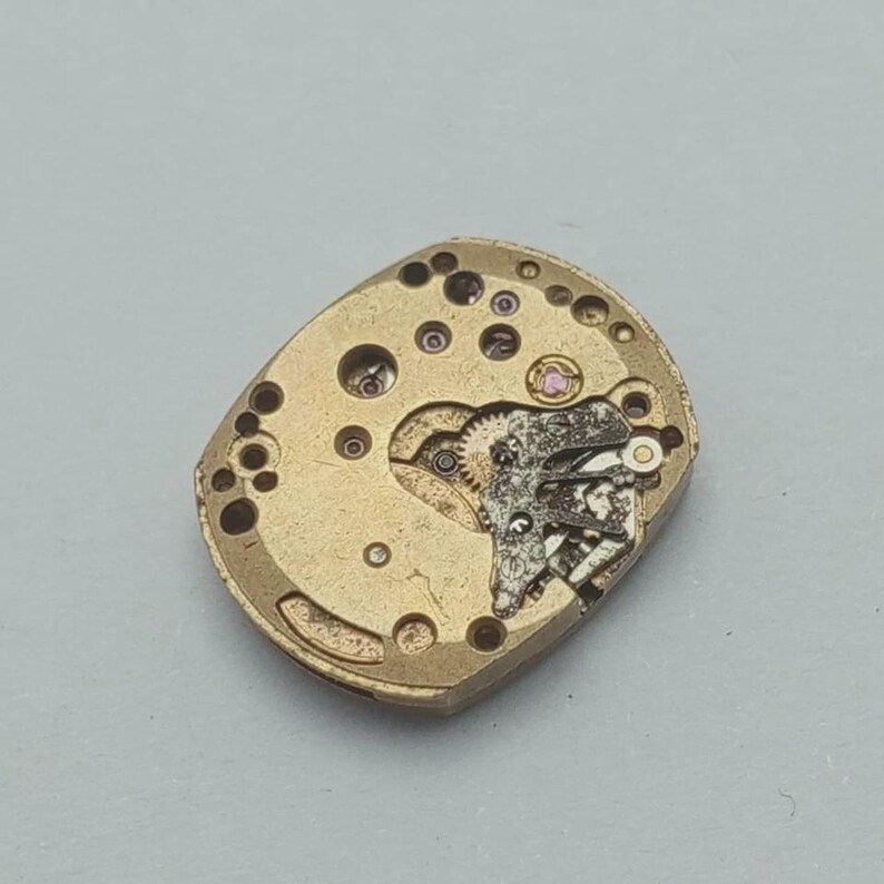 May include: Close-up of a vintage watch movement. The gold-toned mechanism features intricate gears, small jewels, and various components. The movement is oval-shaped with multiple holes and a complex internal structure, showcasing the precision of watchmaking.
