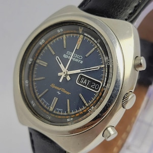 Pode incluir: Um relógio Seiko 5 Sports Speed-Timer vintage com mostrador azul escuro e caixa prateada. O relógio apresenta uma pulseira de couro preta e exibe o dia como "SAT 20". Possui ponteiros e marcadores prateados e uma luneta giratória.
