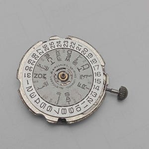 Peut inclure: Un mouvement de montre argent&eacute; avec des inscriptions en arabe et en anglais, incluant les jours de la semaine et les chiffres de 1 &agrave; 31. Le mouvement a une petite couronne et des engrenages internes.