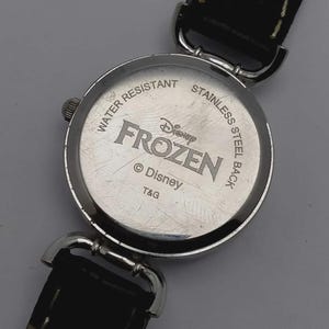 以下が含まれることがあります： 黒い革ストラップが付いた、シルバーカラーの時計の裏側のクローズアップ。時計の文字盤には「FROZEN」の文字とディズニーのロゴが刻まれています。時計には「WATER RESISTANT」と「STAINLESS STEEL BACK」の文字が書かれています。
