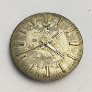 K&ouml;nnte beinhalten: Ein Vintage-Zifferblatt mit einem goldfarbenen, gealterten Aussehen. Das Zifferblatt tr&auml;gt den Markennamen "CAM GENEVA" und das Wort "Popular". Die Zeiger sind silber- und goldfarben, mit rechteckigen Stundenmarkierungen. Die Worte "SWISS MADE" sind ebenfalls sichtbar.