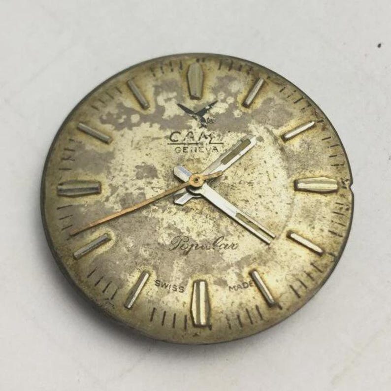 K&ouml;nnte beinhalten: Ein Vintage-Zifferblatt mit einem goldfarbenen, gealterten Aussehen. Das Zifferblatt tr&auml;gt den Markennamen "CAM GENEVA" und das Wort "Popular". Die Zeiger sind silber- und goldfarben, mit rechteckigen Stundenmarkierungen. Die Worte "SWISS MADE" sind ebenfalls sichtbar.